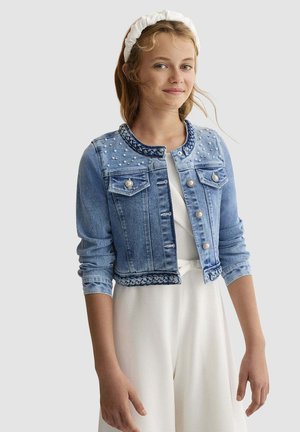 Ragazza giovane che indossa un vestito bianco e una giacca corta di denim blu con dettagli di perle e bordo intrecciato, in piedi davanti a uno sfondo semplice.