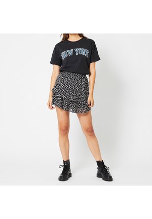 Mujer con camiseta negra con la palabra "New York", minifalda negra con flores y botas negras con cordones hasta el tobillo, posando frente a un fondo blanco.