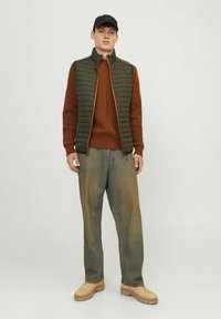 Vesta verde căptușită cu fermoar portocaliu, pulover texturat de culoare ruginită, pantaloni lejeri din denim deschis și pantofi bej cu talpă groasă, purtând o șapcă neagră.