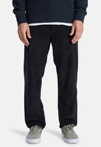 Svarta kordbyxor med avslappnad passform, med vertikala ribbor och en slät textur, i kombination med grå låga sneakers.