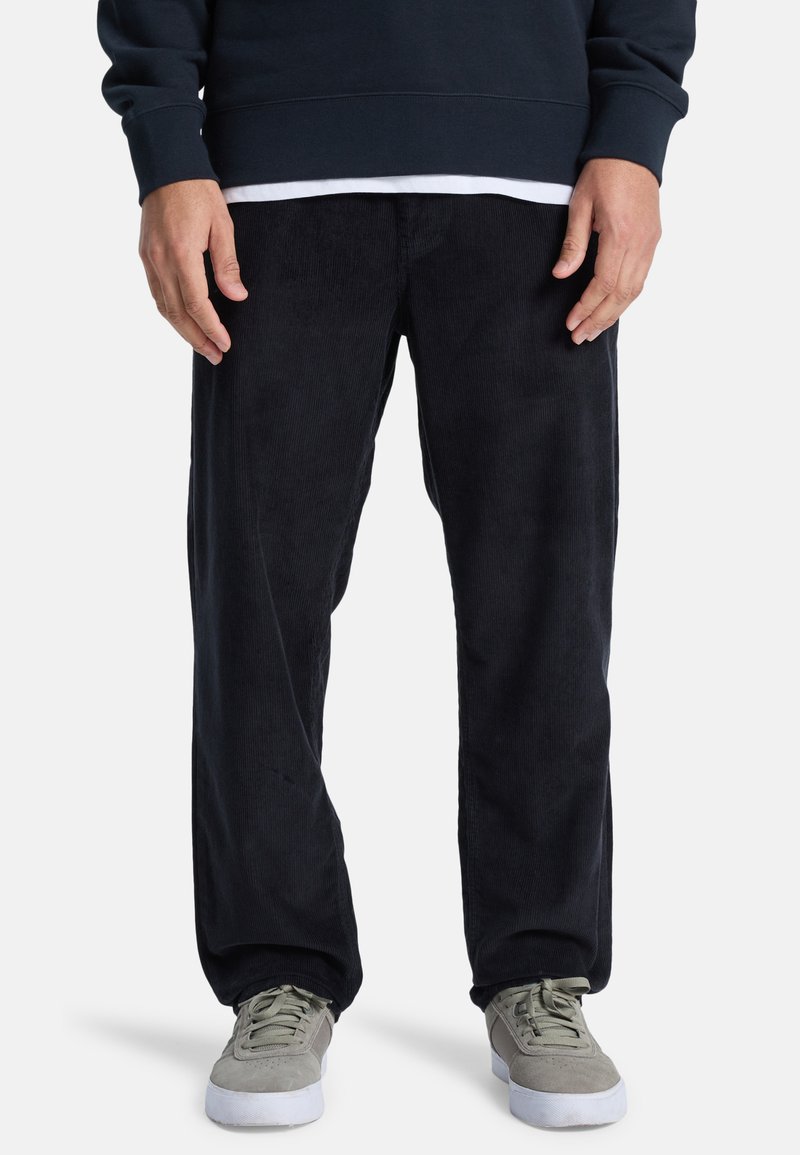 Svarta kordbyxor med avslappnad passform, med vertikala ribbor och en slät textur, i kombination med grå låga sneakers.