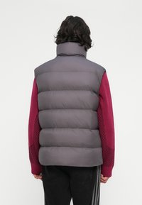 Pessoa com cabelo encaracolado escuro, vestindo um colete acolchoado cinza sobre um suéter de malha vermelho e calças pretas, vista de costas sobre um fundo branco.