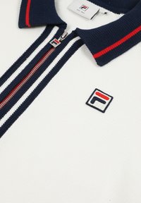 Polo bianca in maglia con colletto a righe blu navy e rosse, zip a righe blu navy e bianche, e piccolo logo FILA sul petto.