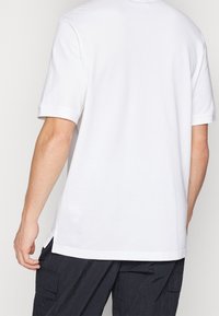 Valkoinen puuvillapaita, jossa on pyöreä kaula-aukko ja lyhyet hihat. Sisältää sivurei'ät helmassa lisäämään liikkuvuutta ja mukavuutta.