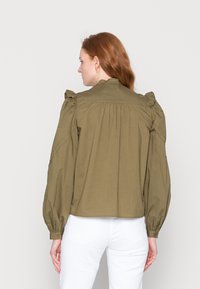 Blusa verde oliva com ombros volumosos, mangas longas e detalhe franzido nas costas. Feita de um tecido leve, possui punhos com botões.