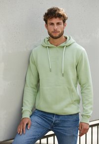 Lichtgroene hoodie met een buidelzak en aantrekkoorden, gemaakt van een textuurstof. Geraadpleegd met blauwe spijkerbroeken.