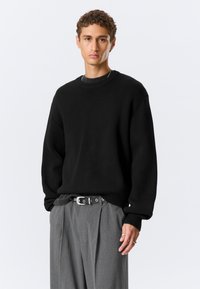 Jeune homme debout, la main gauche dans la poche, portant un pull noir oversize, un pantalon gris à fines rayures, une ceinture noire et des bagues argentées à la main droite.