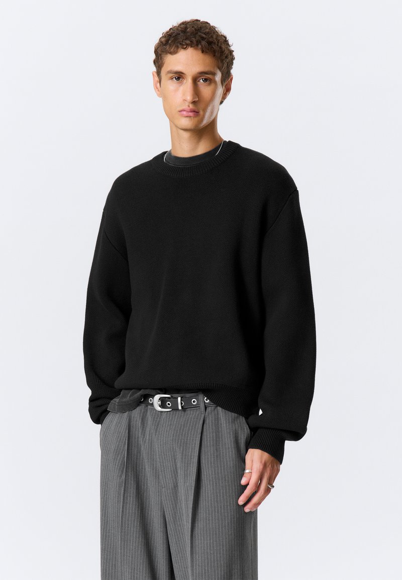 Jeune homme debout, la main gauche dans la poche, portant un pull noir oversize, un pantalon gris à fines rayures, une ceinture noire et des bagues argentées à la main droite.