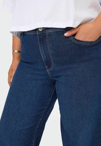 Tumši zili džinsi ar gludu tekstūru, ar priekšējo kabatu, kontrastējošu šuvju un pogu aizdari jostasvietā.