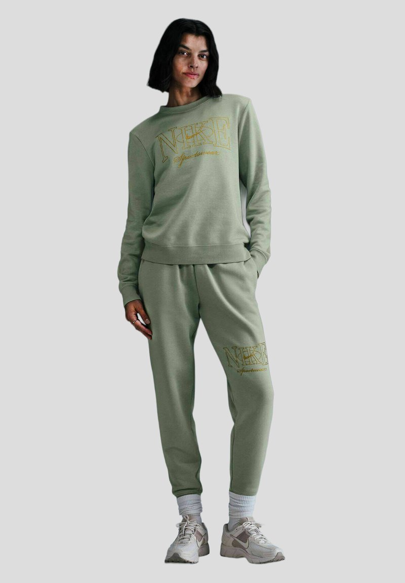 Felpa in cotone verde e pantaloni jogger abbinati con stampa dorata "NIKE". Presenta polsini e caviglie a costine, abbinati a sneaker chiare.
