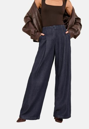 Žena nosí tmavohnedý top so štvorcovým výstrihom, oversized tmavohnedú koženú bundu, široké tmavomodré nohavice a hnedé špicaté topánky.