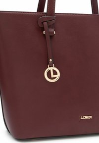 Burgundy leather tote bag with two handles, featuring a gold circular pendant logo and embossed brand name at the bottom. Smooth texture.