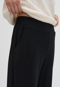 Zwarte broek met een gladde textuur, voorzien van een elastische tailleband en zijzakken. De stof lijkt zacht en iets rekbaar.
