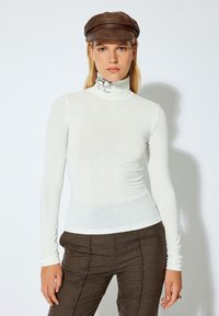 Witte lange mouwen turtleneck gemaakt van een dunne, elastische stof, met een decoratief ontwerp op de kraag, gecombineerd met donkerbruine broeken.