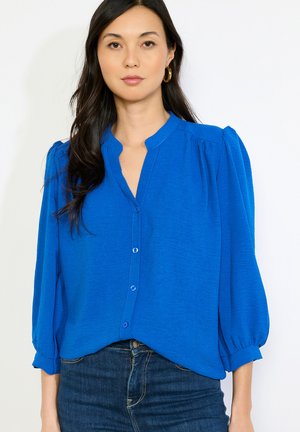 Kobieta o długich ciemnych włosach, nosząca złote kolczyki koła, jasnoniebieską, zapinaną na guziki bluzkę z bufkami oraz ciemnoniebieskie jeansy.