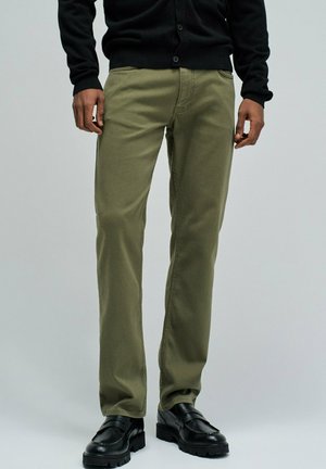 Hombre vistiendo pantalones verde oliva, cárdigan negro y zapatos de cuero negro, de pie contra un fondo liso gris claro.