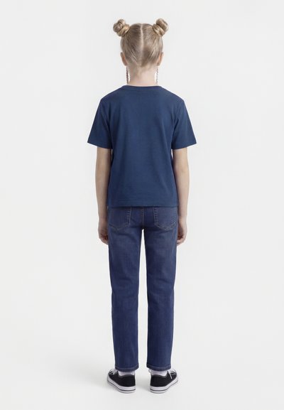 Kind mit blonden Haaren in zwei Dutts, trägt ein dunkelblaues T-Shirt, blaue Jeans, weiße Socken und schwarze Turnschuhe, steht mit dem Rücken zur Kamera.