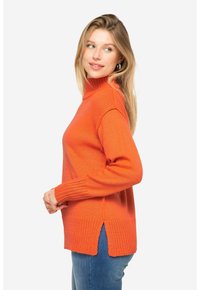Pomarańczowy sweter dziany z wysokim dekoltem, prążkowanymi mankietami, rozcięciami po bokach u dołu oraz teksturowanym wzorem, zestawiony z niebieskimi dżinsami.