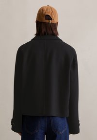 Veste en mélange de laine noire avec un col structuré, des épaules tombantes et des poignets à boutons, agrémentée d'une casquette marron à l'arrière.