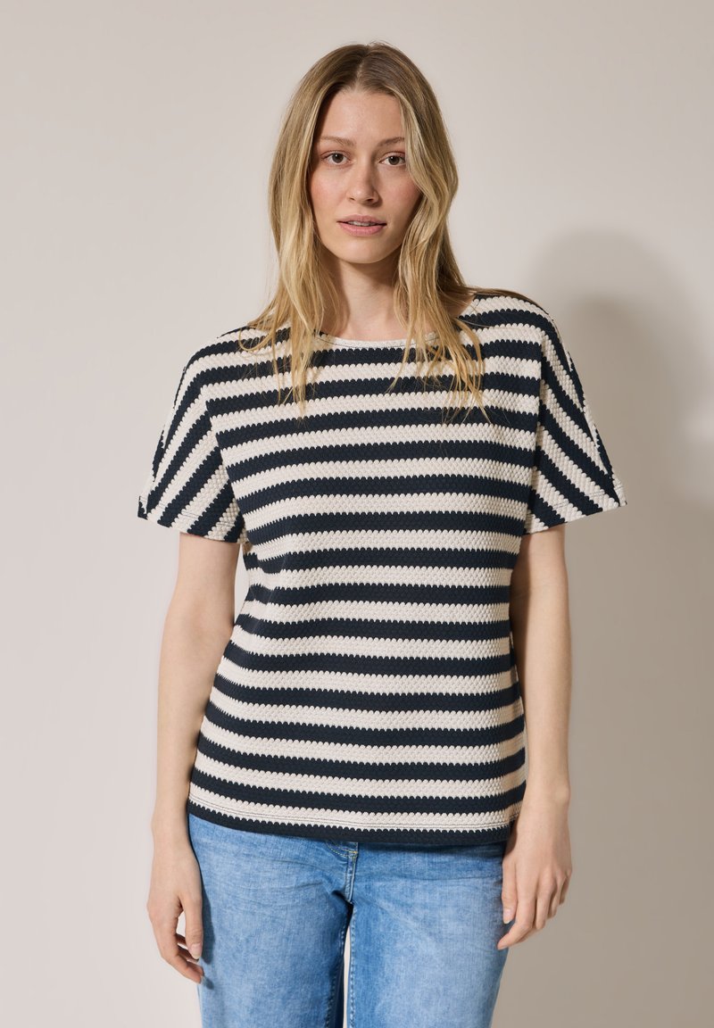 Gestreifter, gestrickter Pullover in Marineblau und Creme, mit kurzen Ärmeln und rundem Halsausschnitt. Weiche Textur und lässige Passform. Getragen mit hellblauen Jeans.