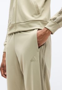 Χακί φόρμα με μαλακό, ελαστικό ύφασμα· τσέπες με φερμουάρ· ριμπ μανσέτες· και τρεις κάθετες ρίγες κατά μήκος των ποδιών του παντελονιού.