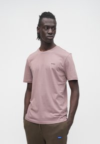 Tricou cu mânecă scurtă și guler rotund, din bumbac roz deschis, cu un mic logo „HUGO” contrastant pe piept. Materialul din bumbac este neted și ușor.