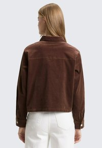 Bruine corduroy jas met een cropped ontwerp, voorzien van een kraag, manchet met omslag en een gestructureerde afwerking. Witte jeans zichtbaar daaronder.