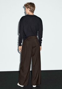 Un pull noir à coupe décontractée, associé à un pantalon large à taille haute marron. Mannequin portant des baskets blanches, se tenant devant un fond uni.