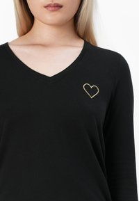 Naine kannab musta V-kaelusega särki, mille vasakul rinnal on tikandina väike kuldne südame kontuur, valge tausta ees.