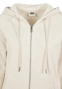 Kremfarget zip-up hettegenser med hette, snor og frontlomme. Laget av myk stoff, med en sølvfarget glidelås og etikett innvendig.
