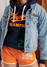 Giacca di jeans con bottoni, sovrapposta a una felpa blu navy con la scritta "Superdry" e "CHAMPIONS" in arancione. Pantaloni corti arancioni con dettagli bianchi.