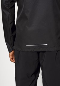 Bakifrån av en person som bär en svart långärmad jacka med en liten horisontell reflexremsa nära den nedre fållen och svarta byxor.