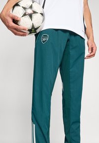 Teal soccer training pants with white side stripes, featuring an Arsenal logo on the left thigh and worn while holding a patterned soccer ball.