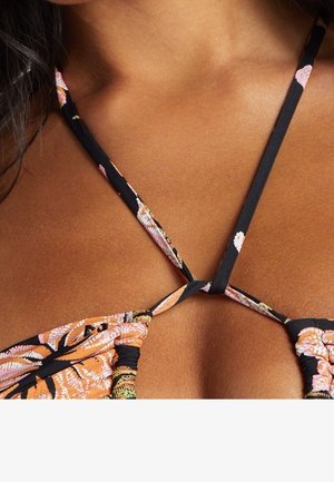 Nahaufnahme der Brust einer Frau, die ein gemustertes Bikinioberteil mit dünnen, schwarzen Blumenbändern trägt, die in der Mitte gebunden sind.
