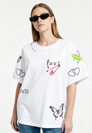 Femme portant un t-shirt blanc oversize avec des motifs de cœurs, papillon, épingle de sûreté et pansement, ainsi que des lunettes de soleil noires.