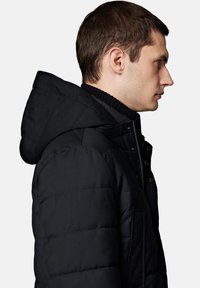 Czarna kurtka puchowa z kapturem, z pikowanym wzorem, przednim zamkiem błyskawicznym, zapięciem na guziki i ściągaczem przy kołnierzu. Widok z boku.
