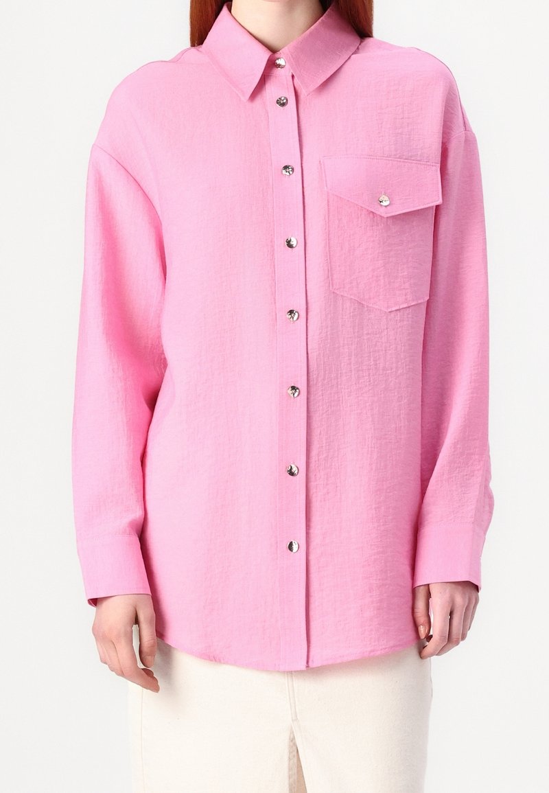 Femme portant une chemise rose à manches longues boutonnée avec une poche poitrine et un pantalon blanc sur un fond uni.