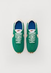 Žalios tinklelio ir zomšinės sportinės kedės su rožinėmis detalėmis, baltomis raišteliais ir mėlynu vidpadžiu. Ant liežuvio ir šonų yra Nike logotipas.