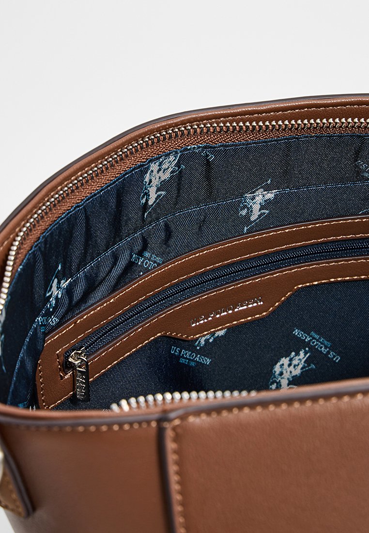 Das Innere der braunen Ledertasche verfügt über ein marineblaues Futter mit Mustern des U.S. Polo Assn. Logos, einer Reißverschlusstasche und strukturierten Details.