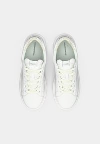 Balta Karl Lagerfeld sporta apavu pāris ar baltām šņorēm, logotips uz mēles un zoles, skatīts no augšas uz baltas fona.