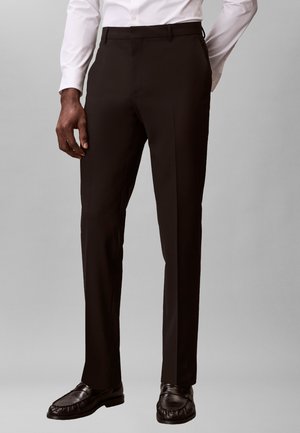 Mann, der schwarze maßgeschneiderte Hosen, ein weißes Hemd und schwarze Leder-Loafers trägt, vor einem schlichten grauen Hintergrund steht.