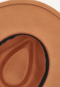 Tan kleurige hoed met een brede rand en een gladde textuur. Heeft een zwarte band langs de binnenrand. Ronde vorm met een naadloze afwerking.