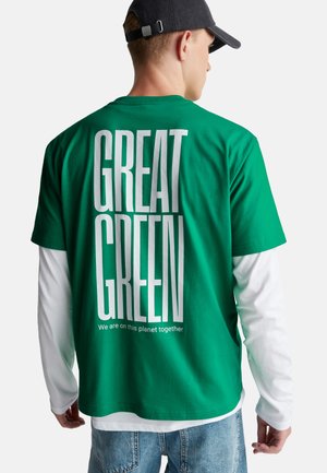 Das grüne Baumwoll-T-Shirt hat auf der Rückseite den auffälligen weißen Text "GREAT GREEN" mit zusätzlichem Text darunter. Es wird über einem weißen Langarmhemd getragen.