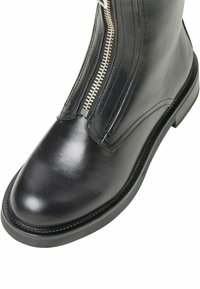 Svart läderankelsko med slät textur, med en silvrig dragkedja och rund tå, placerad på en platt gummisula med sömnadsdetaljer.