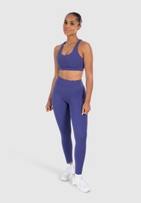 Reggiseno sportivo viola e leggings high-waisted abbinati realizzati in tessuto elastico; il modello indossa scarpe da ginnastica bianche. Texture liscia, branding minimale.