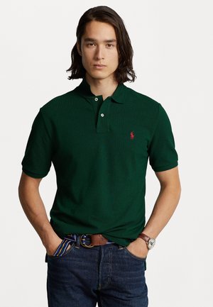 Tricou polo verde cu un guler cu două nasturi, mâneci scurte și un logo roșu pe partea stângă a pieptului, asortat cu blugi albaștri și o curea maronie.