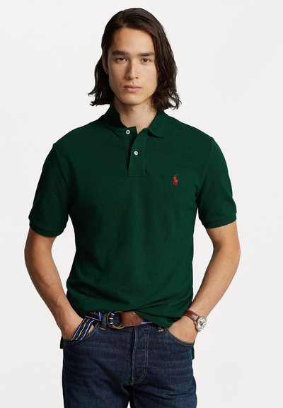 Polo vert avec un plastron à deux boutons, manches courtes et un logo rouge sur la poitrine à gauche, associé à un jean bleu et une ceinture marron.