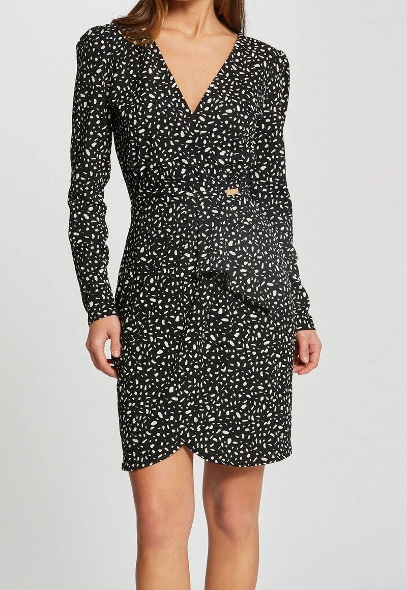 Femme portant une robe portefeuille noire à manches longues avec un motif moucheté blanc et un détail noué à la taille.
