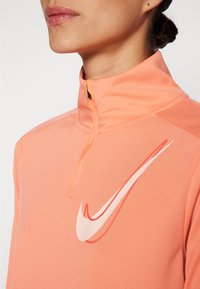 Sportska majica sa patentnim zatvaračem u svijetlo narančastoj boji, s istaknutim bijelim Nike swoosh logom, teksturiranim materijalom i prilagođenim dizajnom.