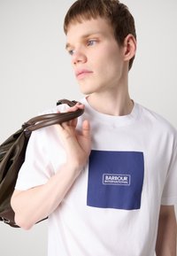 Bílé bavlněné tričko s čtvercovým námořnickým modrým nášivkem se textem "BARBOUR INTERNATIONAL". Model drží hnědý popruh tašky.